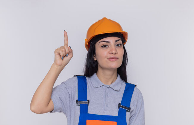 建设者身穿建筑制服 头戴安全帽的年轻建筑工人面带微笑地看着前面 食指站在白色的墙上微笑建筑安全