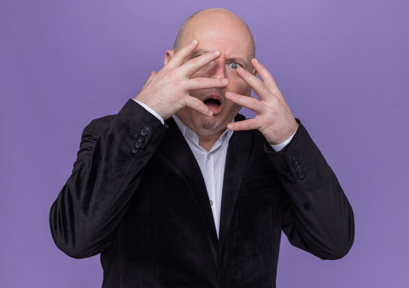 衣服一个穿着西装的中年秃头男人站在紫色的墙上 用手掌遮住脸 用手指看着前面 脸上有疤痕站着手指脸