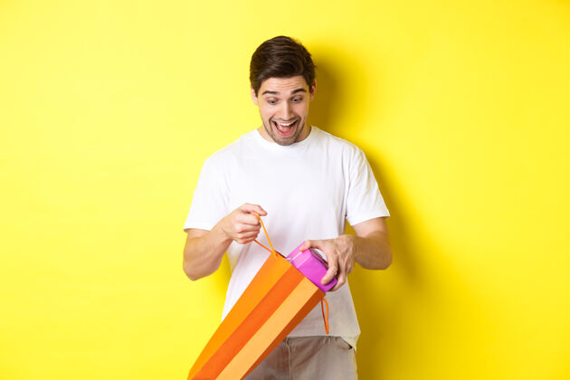 年轻节日和庆祝的概念年轻人站在黄色背景下 惊讶地从购物袋里拿出礼物礼品购物袋成人