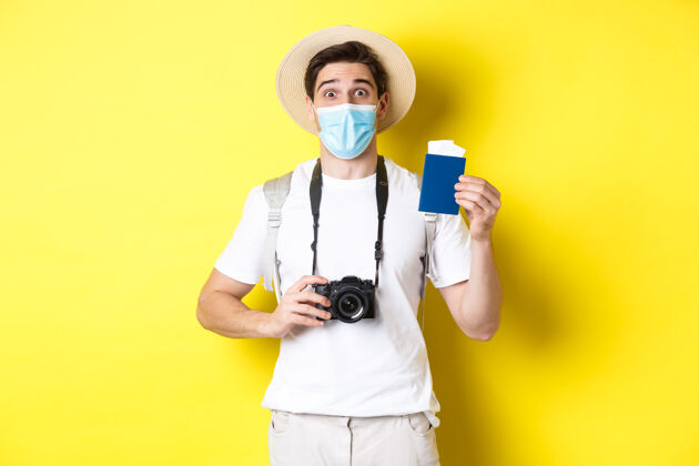 相机covid-19的概念 旅游和检疫带相机的快乐男人游客 出示护照和度假机票 大流行期间旅行 黄色背景模特帅哥站着