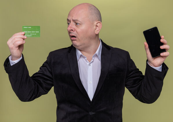 信用中年光头西服男子手持智能手机和信用卡看着它困惑和非常焦虑的站在绿色的墙壁非常困惑移动
