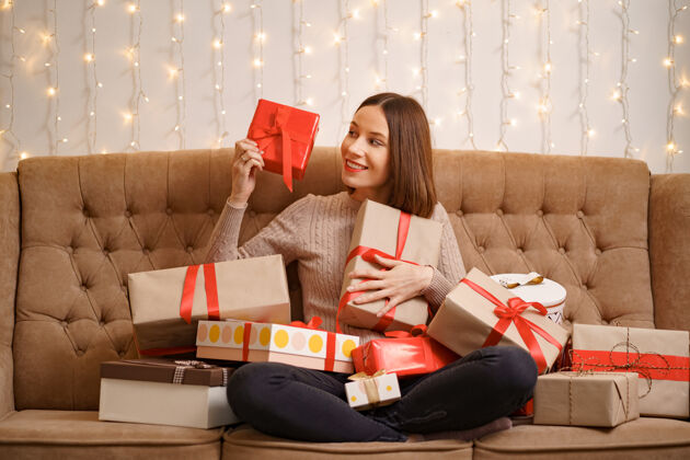 拥抱幸福的年轻女子抱着许多礼物盒盘腿坐在一个驼色沙发与灯幻想礼物毛衣