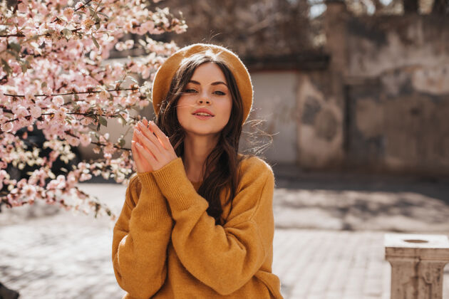 时尚的女人身着橘色毛衣 头戴贝雷帽的迷人女子在樱花身旁合影戴着帽子的黑发卷发女子在阳光明媚的春城漫步太阳镜春天生活