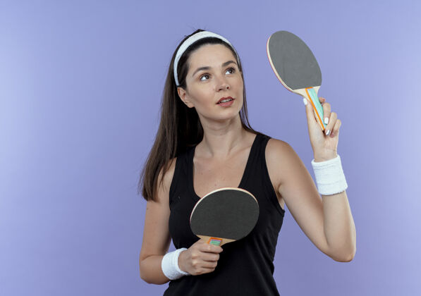 女子戴着头巾拿着球拍准备打乒乓球的年轻健身女士站在蓝色的墙上 神情自信地看着一边持球站立球拍