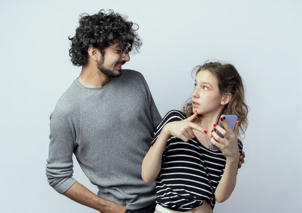 年轻的年轻漂亮的情侣男女 男人看着他迷茫的女友拿着智能手机翻过白墙女人男人抱着