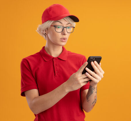 制服身穿红色制服 戴着眼镜 面带严肃表情的年轻送货员站在橙色的墙上看着自己的智能手机帽子穿女人