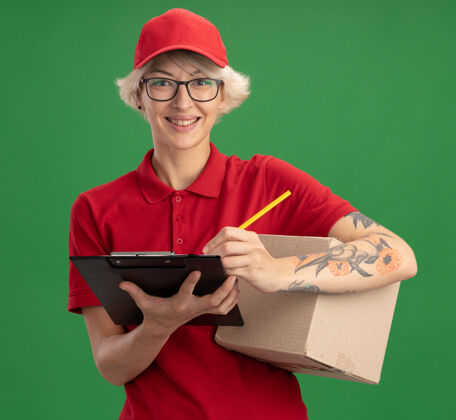 翻页年轻的送货员身穿红色制服 戴着帽子 戴着眼镜 拿着硬纸盒 拿着空白页的剪贴板和铅笔 微笑着站在绿色的墙上写着什么剪贴板什么的拿着