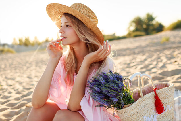 自然浪漫的白人女子 戴着时髦的帽子 穿着优雅的粉色连衣裙 手持草包和花束 在沙滩上摆着姿势年轻好看漂亮