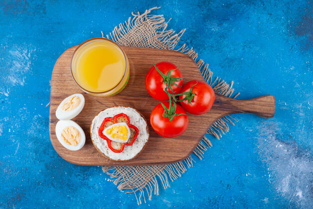 风味芝士面包 一杯果汁 鸡蛋片和整个西红柿放在切菜板上 蓝色的布料上面粉盐番茄