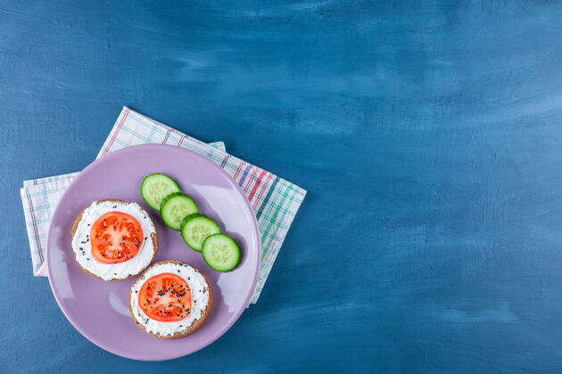 风味西红柿片放在奶酪面包上 黄瓜放在盘子里 茶巾上 蓝色的盘子美味美味