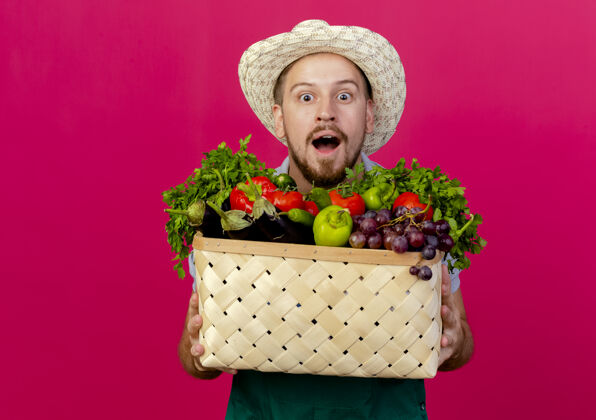 年轻印象深刻的年轻英俊的斯拉夫园丁在制服和帽子举行的蔬菜篮子隔离在深红色的墙壁与复制空间看着帽子篮子