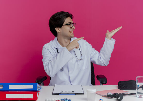 眼镜面带微笑的年轻男性医生 穿着医用长袍 戴着听诊器 戴着眼镜 坐在办公桌旁 手持医疗工具 头对侧地转动 空手指向隔离在粉红色墙上的一侧坐着指着微笑