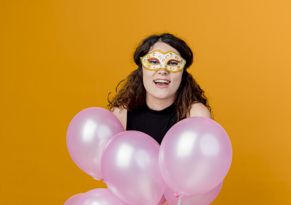 看着年轻漂亮的女人卷发手持一束气球在派对面具快乐快乐的生日派对概念超过橙色生日派对束