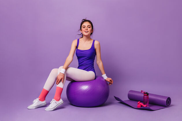 性感穿着明亮健身服的女士坐在瑜伽垫 粉色水瓶和哑铃墙上的健身球上满意移动运动员