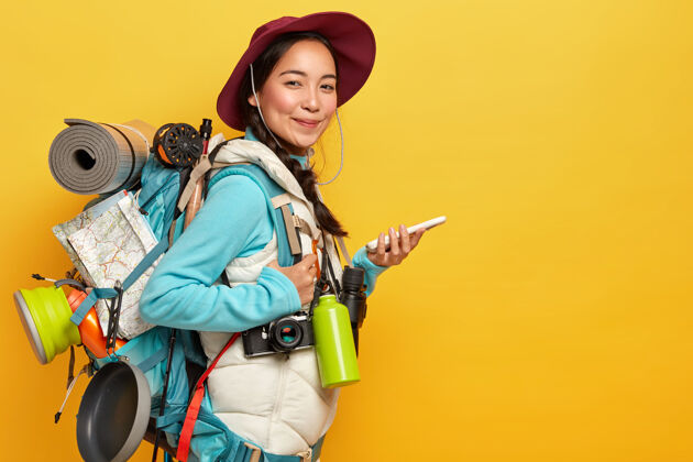 相机亚洲女士表情愉快 用在线导航地图寻找路线 手持手机 戴帽子 休闲服 背背包 烧瓶 望远镜 隔离在黄墙上青年高兴科技