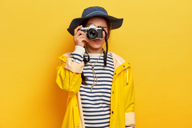 爱好女旅行者在旅途中拍摄难忘的照片 手持复古相机 拍摄美丽的风景或地方 穿着条纹套头衫 雨衣和帽子 隔离在黄色的墙壁上穿民族亚洲人