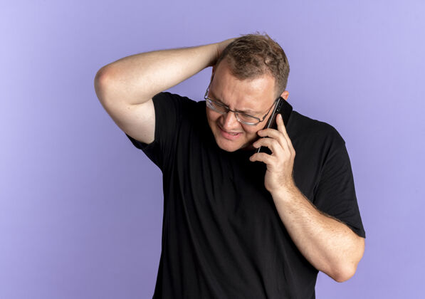 超重戴眼镜的超重男子身穿黑色t恤 一边用蓝色手机交谈 一边显得困惑和不快看困惑电话