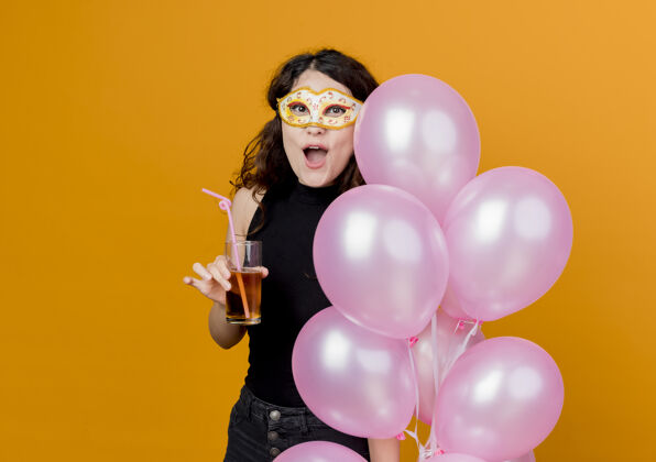 空气年轻漂亮的女士 卷发 手持一束气球和鸡尾酒 戴着派对面具 快乐快乐的生日派对理念 橙色卷曲美丽气球
