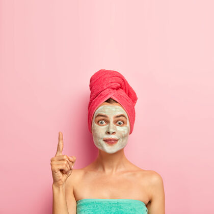 清洁可爱的女人把食指指向上方 戴上面膜 减少粉刺 避免皮肤干燥的问题 用浴巾包着治疗微笑赤膊上阵