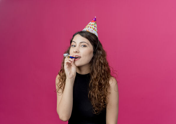 哨子年轻漂亮的女人卷发在节日帽子吹哨子快乐和积极的生日派对概念站在粉红色的墙上卷发年轻积极
