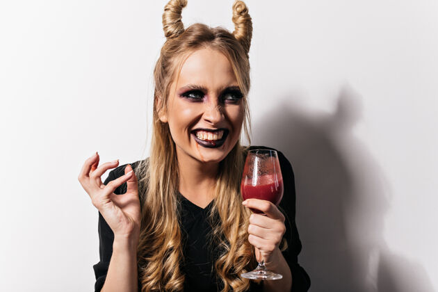 服装一位身着吸血鬼服装的微笑迷人的女士正在享受派对酒杯里装着假血的笑女孩的照片神秘狂欢节神秘