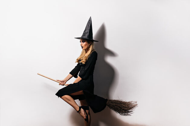 女巫在万圣节 梦幻般的年轻女巫骑着扫帚飞翔室内拍摄的温文尔雅的金发巫师在白墙上摆姿势巫师幻想帽子
