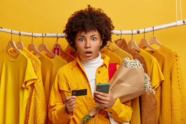 非洲便利的银行和网上购物概念惊呆了的年轻非洲裔美国女士惊讶地盯着相机 拿着手机和花束 背景是挂在衣架上的黄色衣服卡片持有黑色