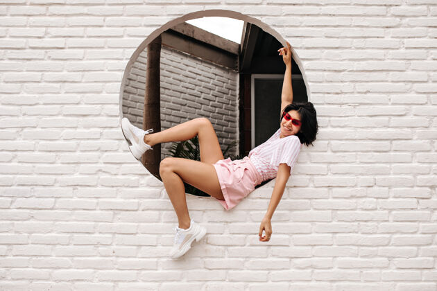 晒黑满意的女人坐在砖砌的墙上 看着相机一个戴着粉色太阳镜和短裤的快乐的女模特的户外镜头生活方式女人装束