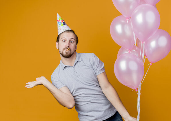 举行戴着节日帽的年轻人举着一堆气球站在橙色的墙上庆祝生日看年轻惊人
