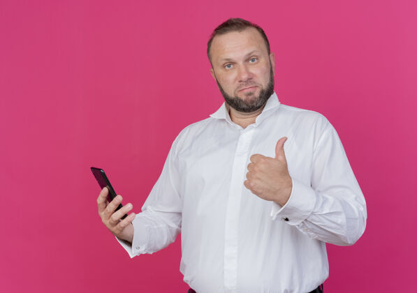 看一个留胡子的男人穿着白衬衫 举着智能手机 竖起大拇指站在粉色的墙上男人站着拇指