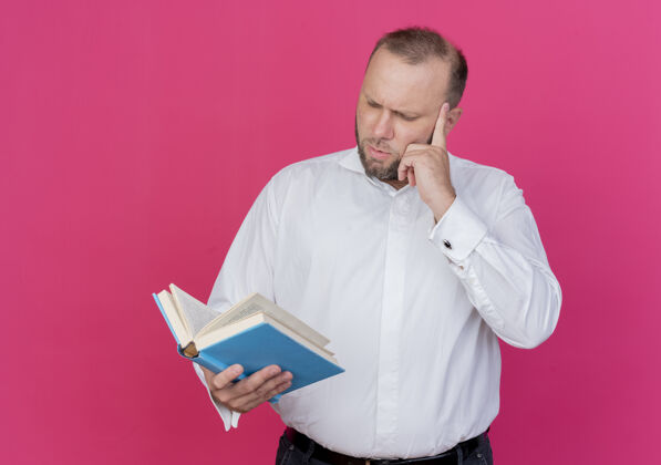 脸一个留着胡子的男人穿着白衬衫 拿着一本打开的书 严肃地站在粉红色的墙上看着它胡子穿开
