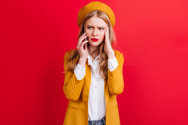 女性一个穿着黄色贝雷帽的金发女人在讲电话穿夹克的白人女孩拿着智能手机站在红墙上室内时尚手机