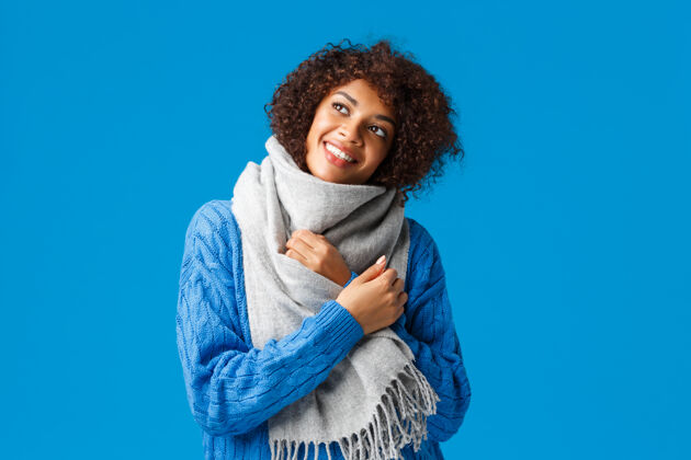 时尚梦幻浪漫无忧无虑的非洲裔美国女人在冬天穿毛衣 戴围巾 抬头沉思 蓝色的墙壁欢乐休闲寒冷