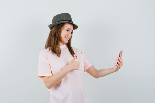 微笑穿着粉色t恤 戴着帽子 看上去很快乐的年轻女孩在视频聊天中竖起大拇指 正面视图童年快乐秀