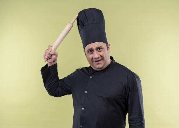 刮男厨师身穿黑色制服 头戴厨师帽 站在绿色背景下 用罗琳别针搔头 看上去很困惑烹饪绿色头