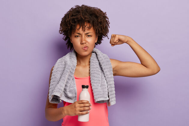 训练锻炼和运动概念健康的运动女性举起手臂 露出肱二头肌 穿着休闲的粉色上衣 拿着一瓶水 脖子上有毛巾擦汗 在室内锻炼二头肌人美国人