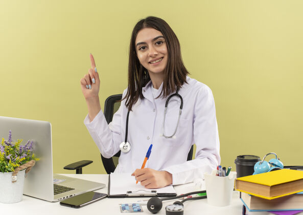 新的年轻的女医生穿着白大褂 脖子上戴着听诊器 食指有了新的想法坐在桌边 笔记本电脑放在墙上女性桌子外套