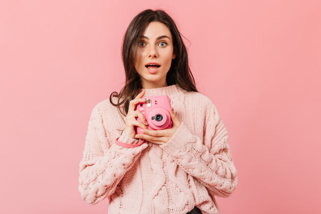 深色一位表情惊异的灰眼睛女士在一台粉色迷你相机上拍照一位深色直发 穿着针织毛衣的女士在一个孤立的背景上摆姿势肖像微笑衣服
