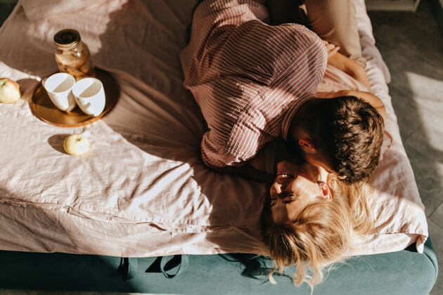 约会一对年轻夫妇在床上闲逛吃早餐穿条纹衬衫的家伙拥抱着笑着的女孩微笑茶欢呼