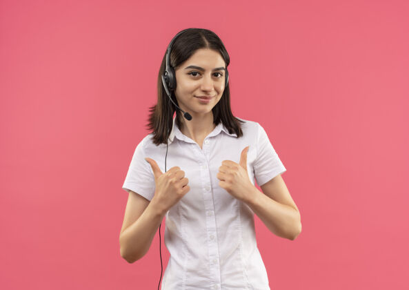 人穿着白衬衫 戴着耳机的年轻女孩 微笑着站在粉色的墙上 竖起大拇指望着前面微笑手势公民