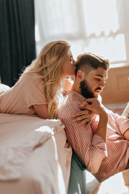 女朋友在明亮的卧室里 卷曲的金发女孩亲吻坐在地板上穿着条纹衬衫的男友成人公寓团聚