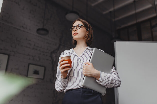 铅笔穿着时髦衬衫和黑色裤子的严肃的女商人在办公室里拿着笔记本电脑喝咖啡科技工作杯子