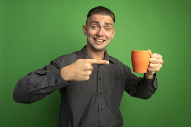 面部表情身穿灰色衬衫的年轻帅哥拿着橙色的马克杯 用食指指着杯子 站在绿色的墙上 开心地笑着人微笑衬衫