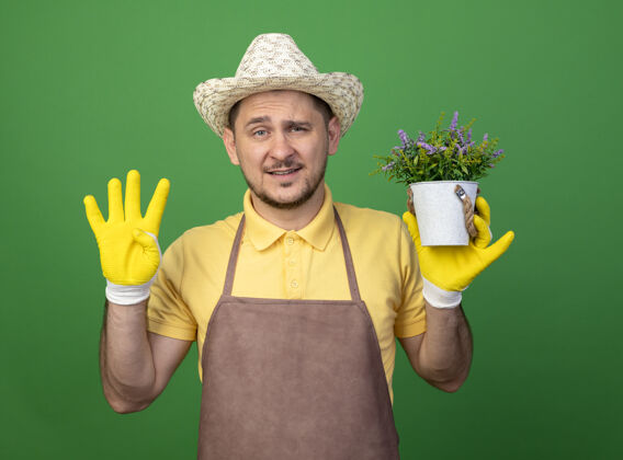 花园年轻的园丁穿着连体衣 戴着帽子 戴着工作手套 手里拿着盆栽植物 看着前面 四号站在绿色的墙上工人人人