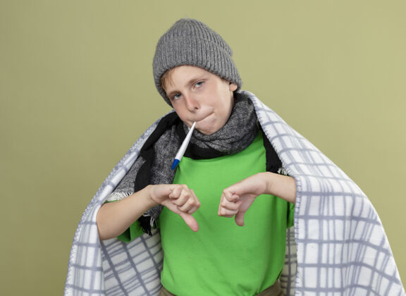 放生病的小男孩穿着绿色t恤 戴着暖和的围巾 帽子裹着毯子 把温度计放进嘴里 测量温度 拇指朝下站在明亮的墙上羽绒服小展示