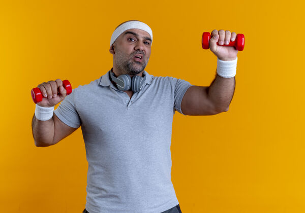 姿势成熟的运动型男子戴着头巾 站在橙色的墙3上练习哑铃 看起来紧张而自信头带人人