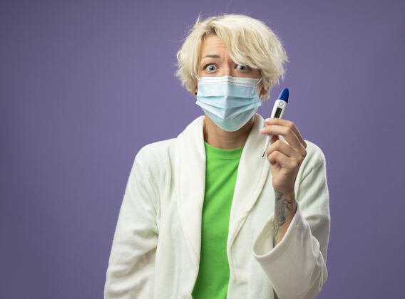 不健病的不健康的女人 戴着护面面具的短发 手里拿着体温计 心烦意乱地站在紫色的墙上女人体温计抱着