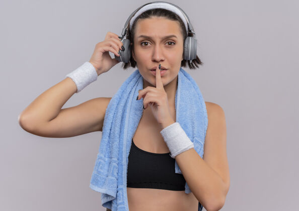 健身一位身穿运动服的年轻健身女士头戴耳机 脖子上套着毛巾 手指放在嘴唇上 站在白色的墙上做着沉默的手势运动公民头