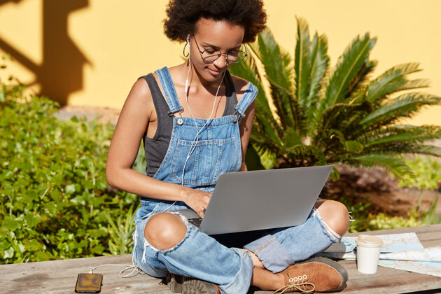 阿特拉斯照片中有一个漂亮的女人 留着非洲发型 盘腿坐着 手提电脑 键盘 博客的新出版物 听音乐 用手机 耳机 穿着时髦的衣服现代地图博客
