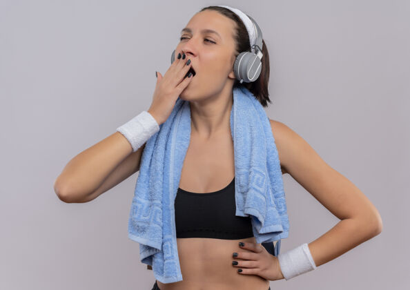 市民一位身穿运动服 头戴耳机 脖子上套着毛巾的年轻健身女士站在白墙上打呵欠 累得想睡觉运动毛巾运动装